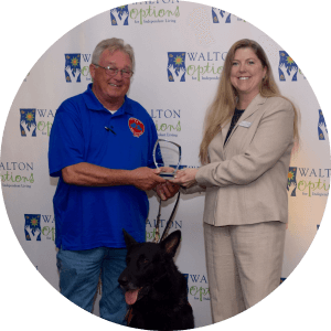 A winner of the MVP Community Awards 2016 is presented with his award by a Walton Options team member in front of the Walton Options Backdrop. The winner also has his service dog next to him.
