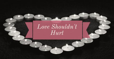 A series of tea light candles are arranged in the shape of a heart and are lit. over the center of them is a pink text banner with the text: Love Shouldn't Hurt