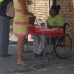 Visitors check in with Shelia - a woman in a yellow and pink dress stands on one side of the table. Shelia is in her wheelchair on the other side of the table speaking to the visitor.