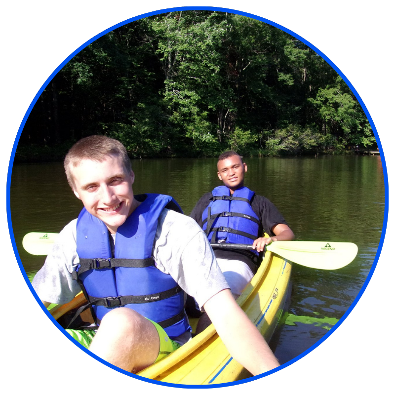 Image is a circle shape with a blue border. Background is a lake with trees. In the foreground are two males wearing life jackets sitting in a canoe posing for a selfie style photo.