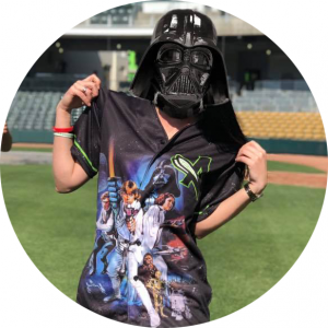 Photo: a person standing on a baseball field showing off a baseball Jersey with the Augusta GreenJackets logo and Star Wars Characters. The person is wearing a Darth Vader Mask.