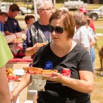 A woman holds a plate with pizza slices, a can of soda and a bag of chips. She is wearing sunglasses as she looks at the snack table during the ADA25 Family Fun Day.