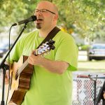 A candid shot of a man in a green ADA25 shirt, playing the guitar and singing into a microphone.