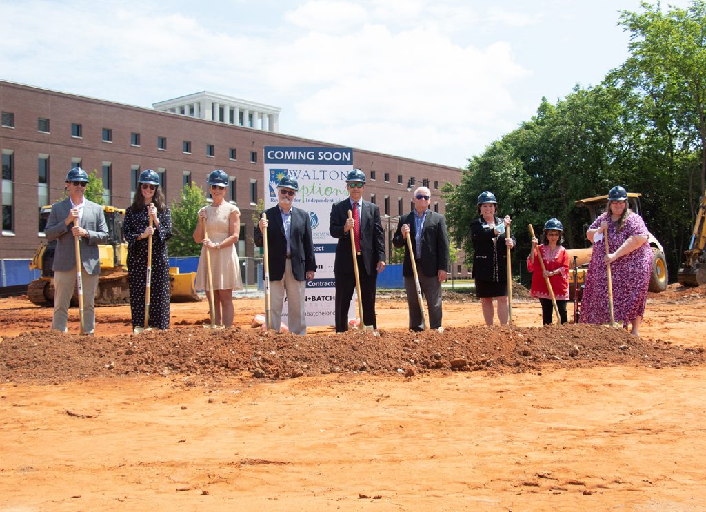 Nine people (five women and four men) stand on a construction lot, of dirt. Each person, wearing a construction hat and holding a shovel, smile toward the camera. A small mound of dirt sits before their feet.